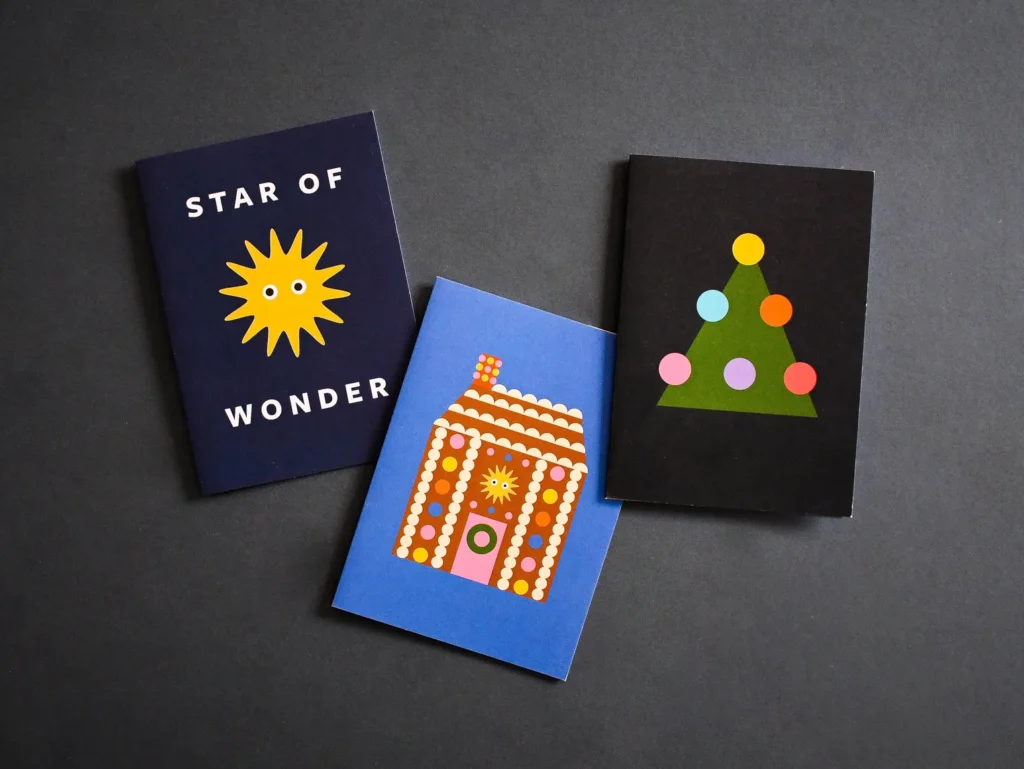 Three Christmas cards standing on a dark grey surface: a navy card reading "Star of Wonder" with a yellow sun/star illustration, a blue card with a gingerbread house design, and a black card with a green Christmas tree made of geometric shapes and coloured baubles.