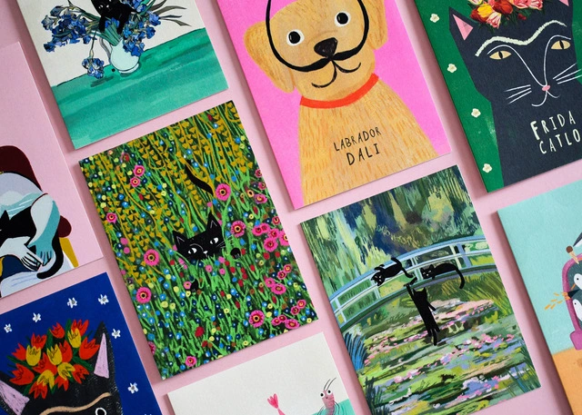 A top-down arrangement of colorful illustrated greeting cards featuring art-themed animals, including a black cat as Frida Kahlo, a dog as Salvador Dalí, and cats in a Monet water lily garden, set on a pink background.