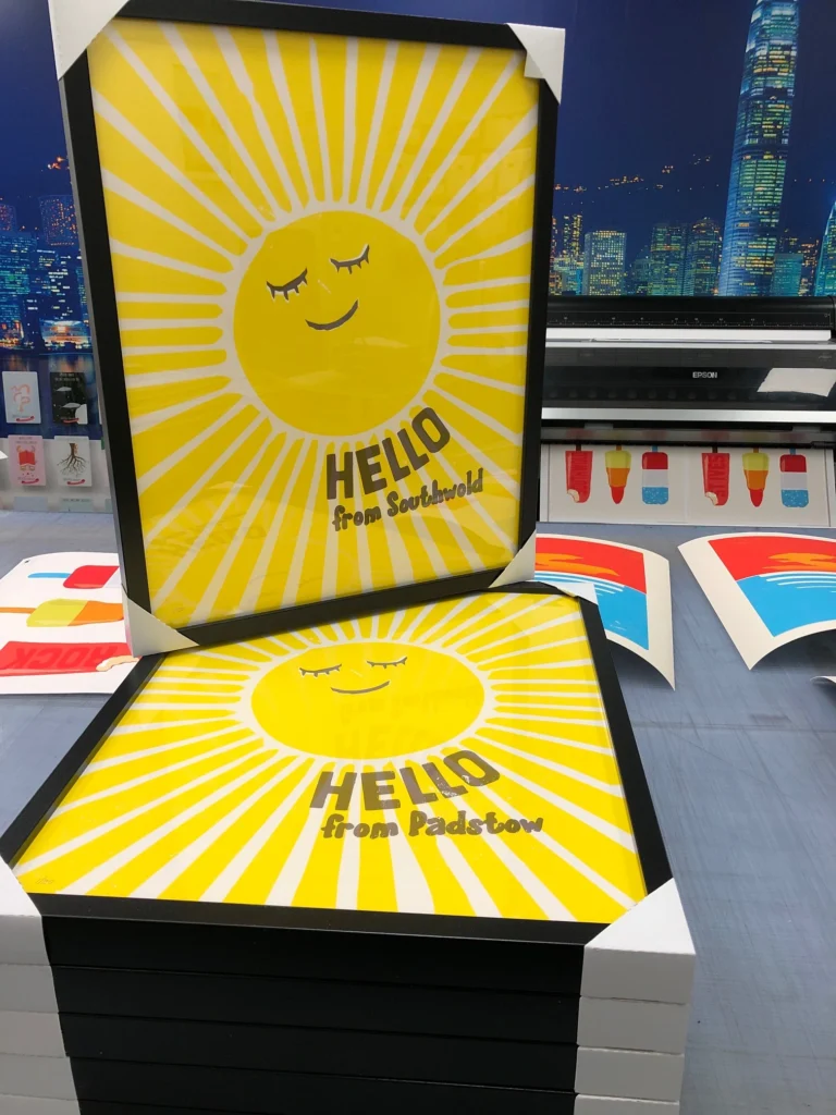 Stacked black-framed art prints with protective corner guards, featuring bright yellow smiling sun illustrations with text reading 'HELLO from Southwold' and 'HELLO from Padstow.'