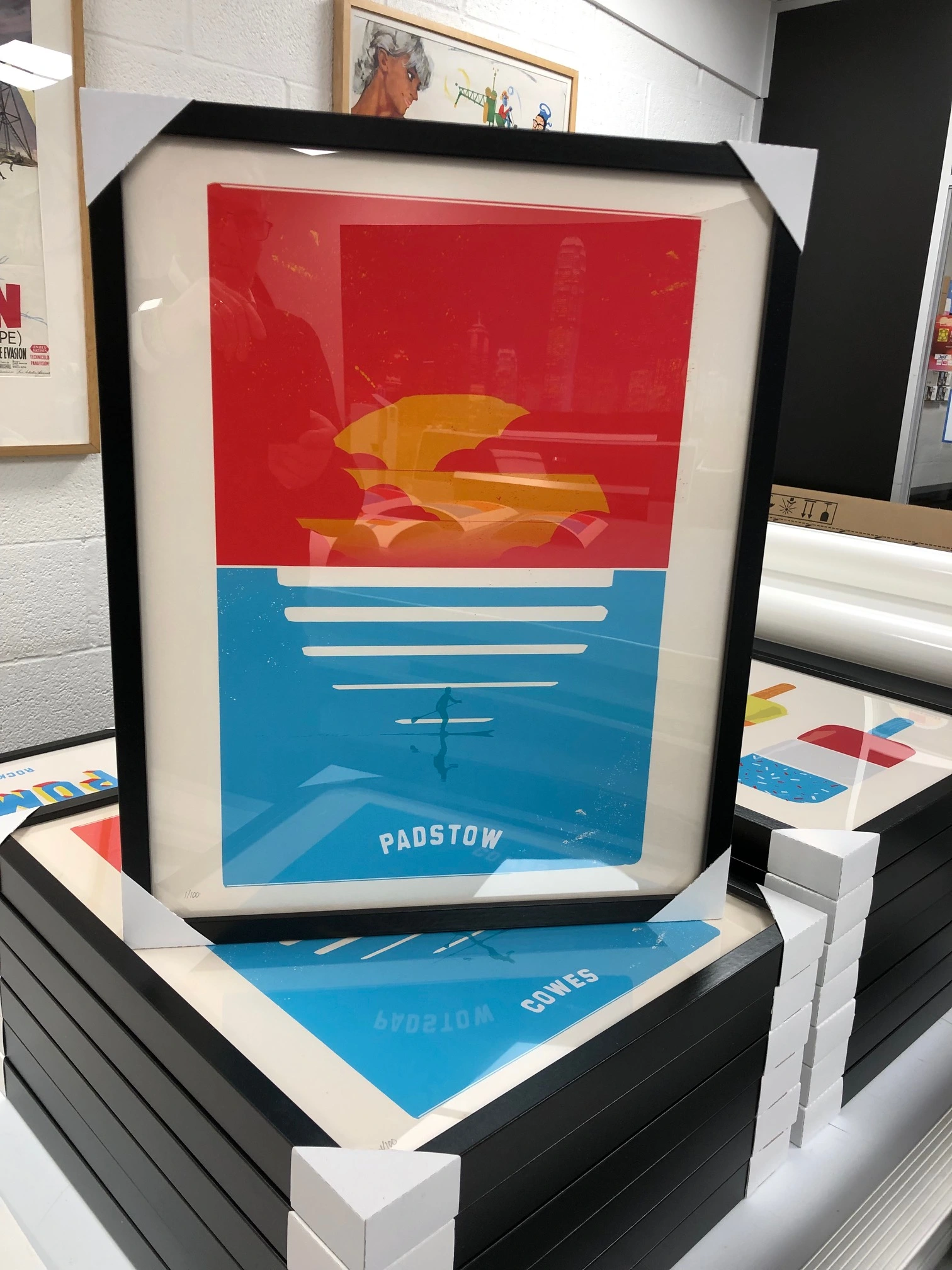 Stack of black-framed art prints with protective corners, showcasing a bold graphic travel poster of Padstow with a red sky and blue water featuring a paddleboarder.
