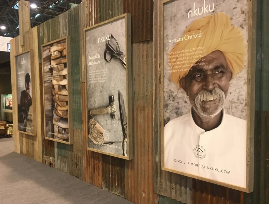 A trade show exhibition wall made of rustic wood and corrugated metal, displaying large framed Nkuku posters celebrating artisan craftsmanship and handmade goods.