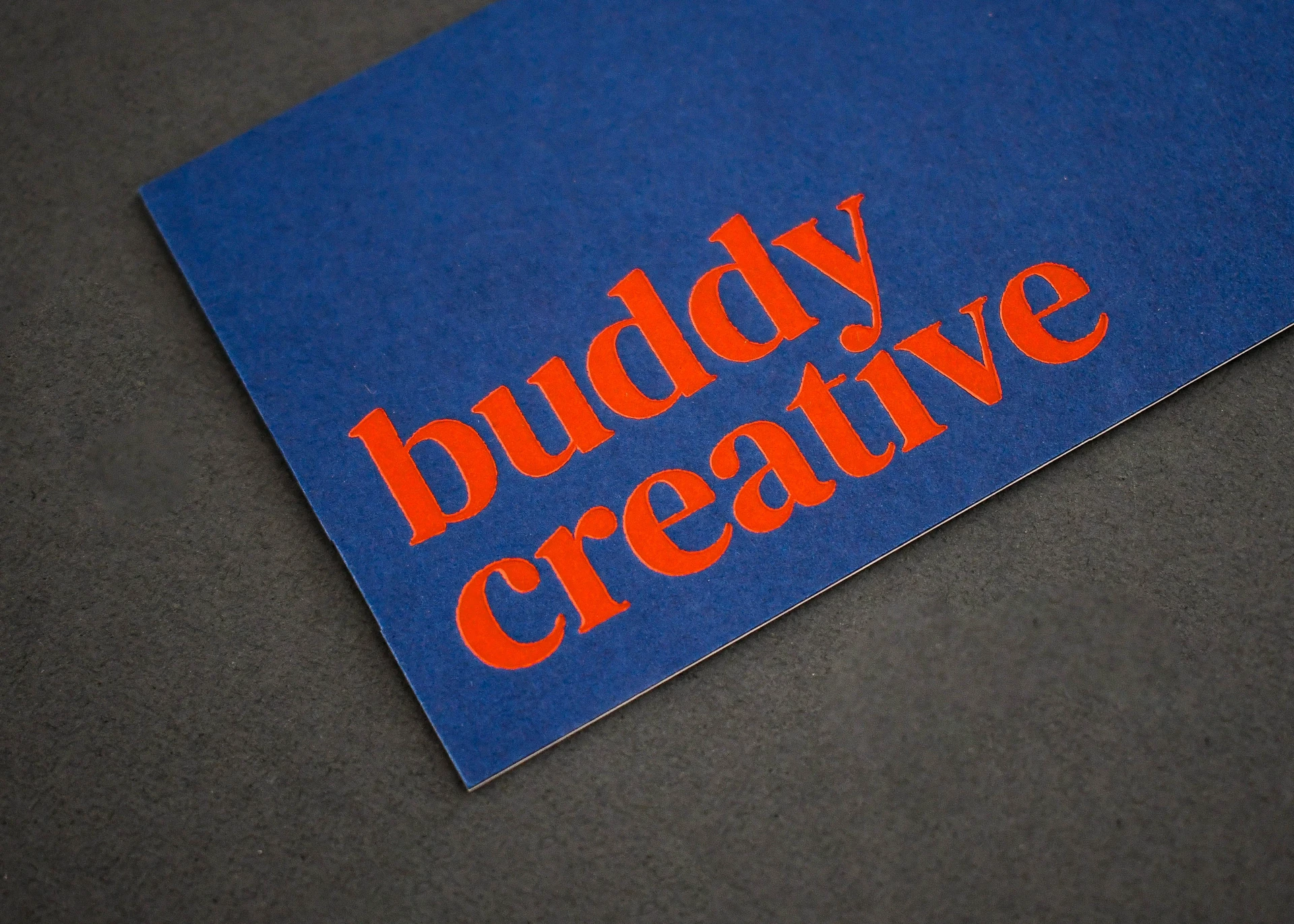 Close-up of a navy "buddy creative" business card showing the deep orange deboss texture.