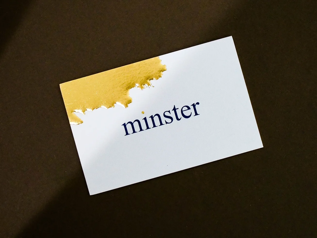 White business card reading "minster" below a gold foil-stamped corner.