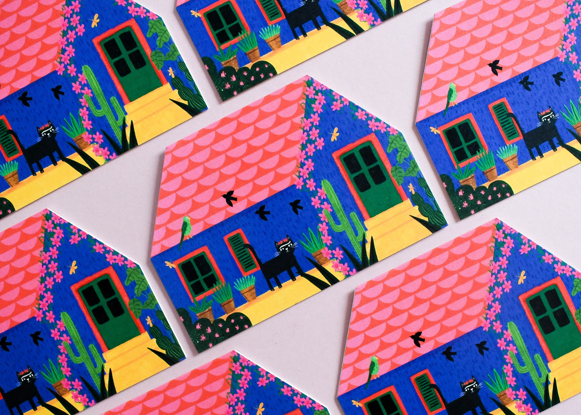 A repeating flat lay of house-shaped die-cut greeting cards illustrating a bright blue cottage with a pink scalloped roof, cacti, and a black cat.