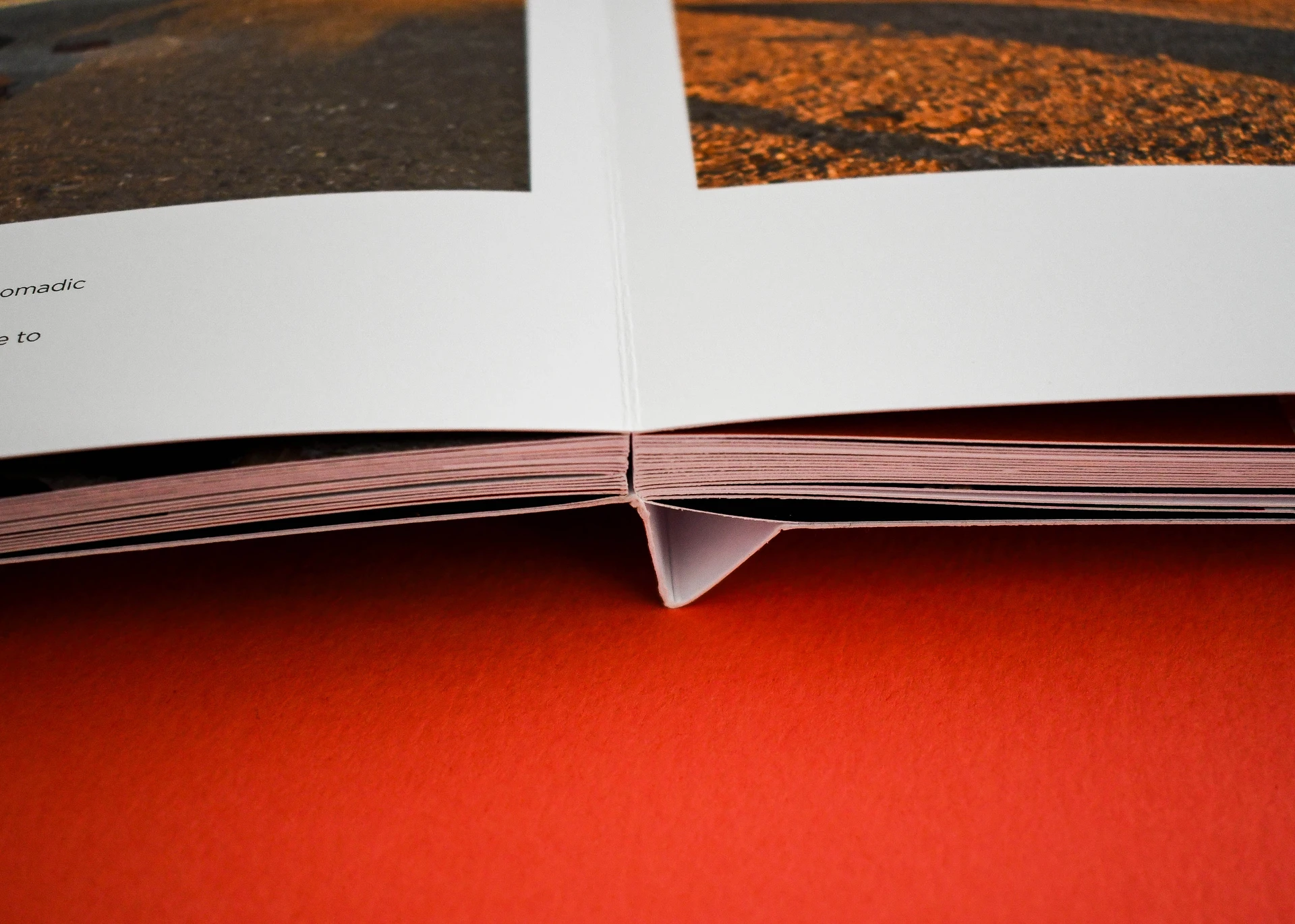 Close-up view of an open lay-flat book on an orange background, showing the special hinge spine construction that allows pages to lay completely flat.