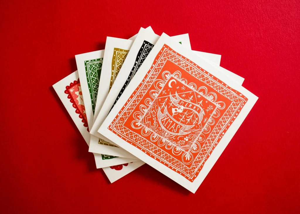 Five square lino-cut style cards fanned out, each with a Merry Xmas border design.