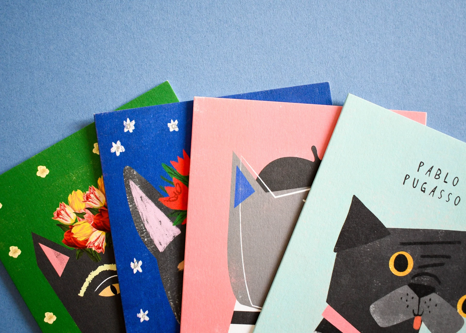 Fanned-out greeting cards on a blue background featuring funny artist-inspired pets, including a pug titled 'Pablo Pugasso' and a cat as Frida Kahlo.