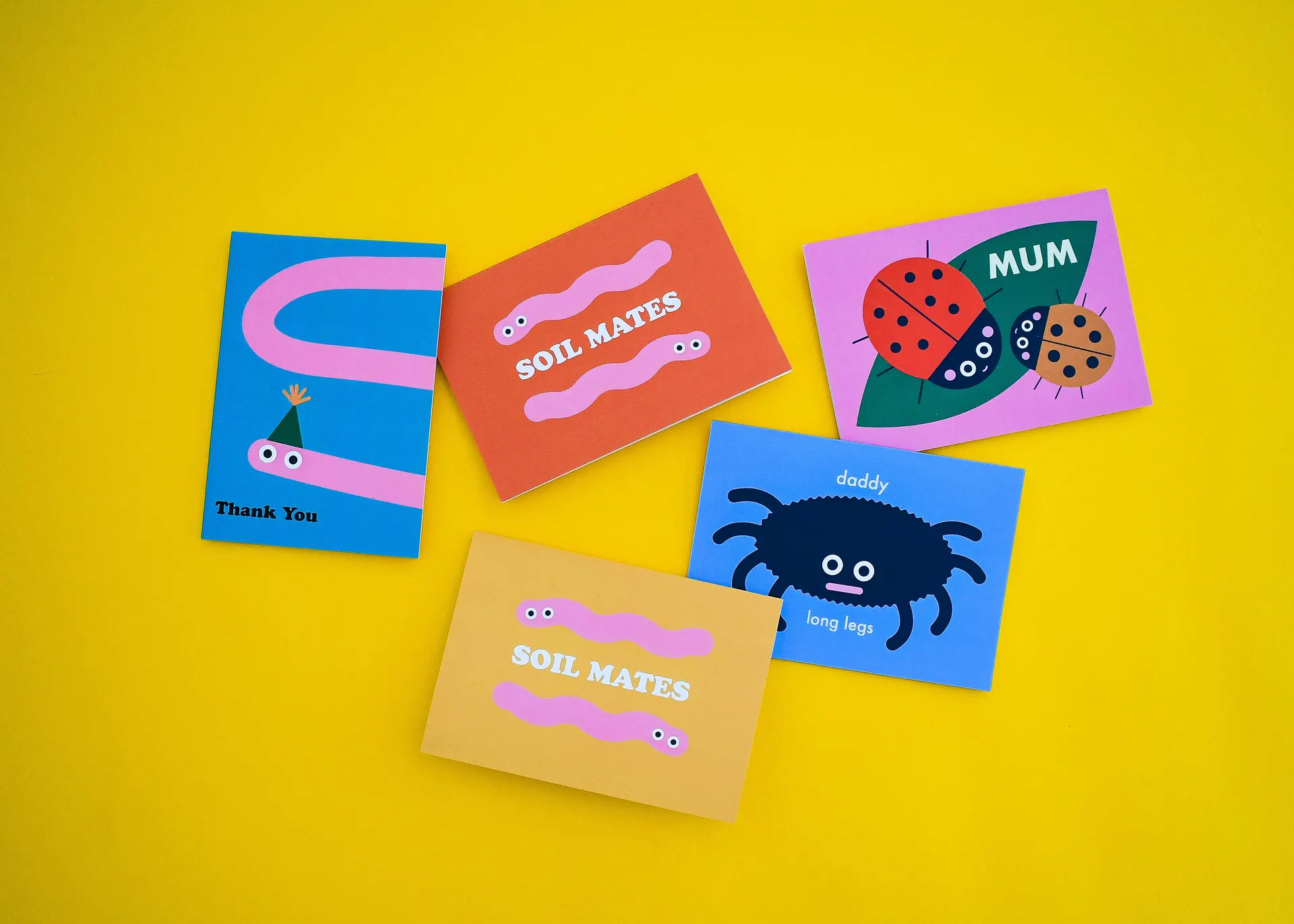 A flat lay of five colorful greeting cards on a bright yellow background featuring quirky bug illustrations, including worm cards reading 'SOIL MATES', a ladybird card for 'MUM', and a 'daddy long legs' spider card.
