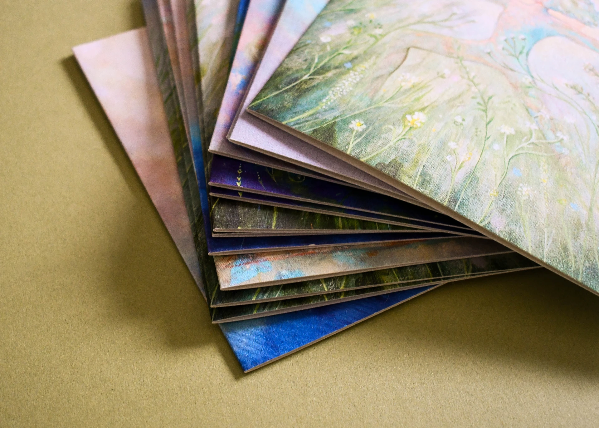 A close-up side view of a stacked, fanned pile of thick paper greeting cards showcasing soft floral and meadow landscape artwork.