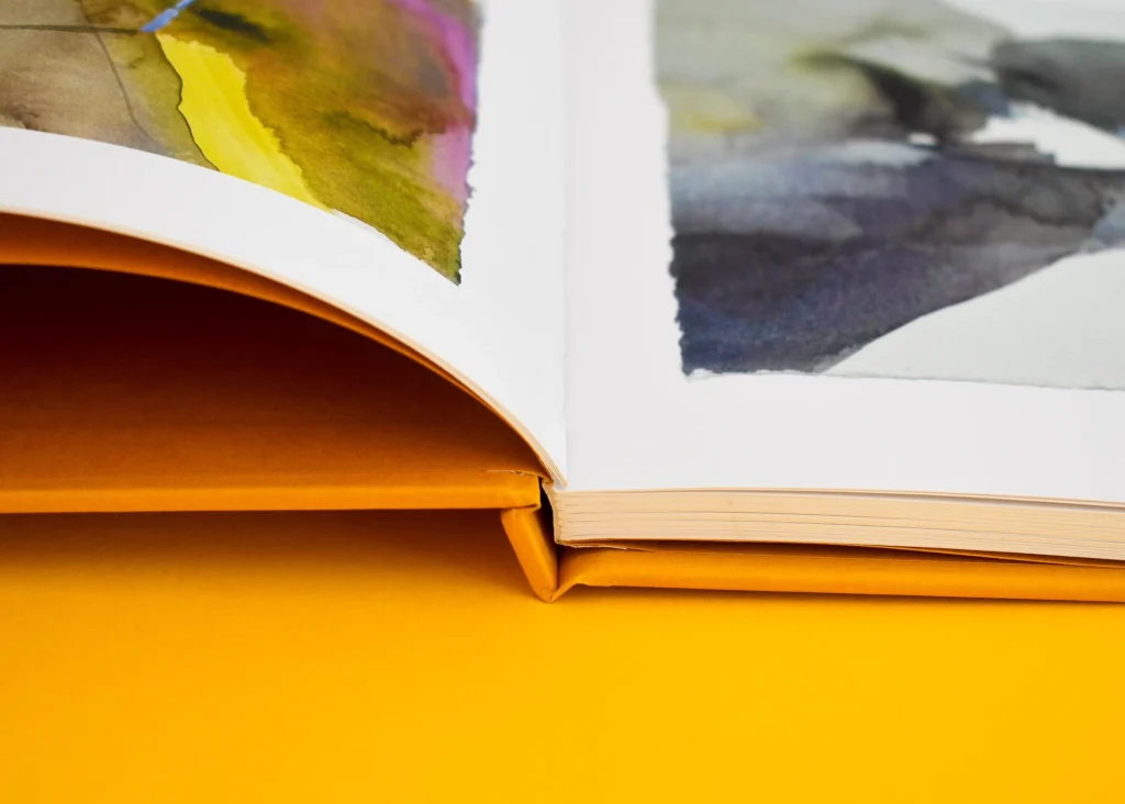 Close-up shot of an open yellow cloth-bound book displaying watercolor artwork and the seamless lay-flat spine construction.